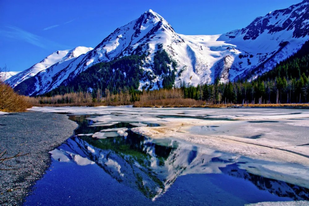 Partially Frozen Lake with Mountain Range Reflected in the Great Alaskan Wilderness. A Beautiful Landscape of Blue Sky, Trees, Rock, Snow, Water and Ice. Near Seward highway near Anchorage, Alaska.